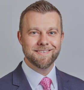 Man smiling at camera with white shirt, light color tie and dark suit. He has a beard and mustache