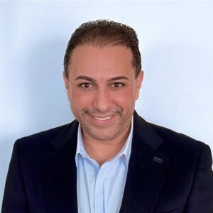 Man in dark suit jacket smiling at the camera with a light blue shirt.